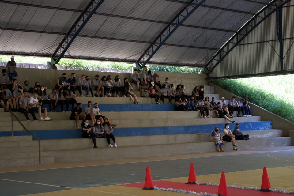 Estudantes sentados na arquibancada da quadra coberta.
