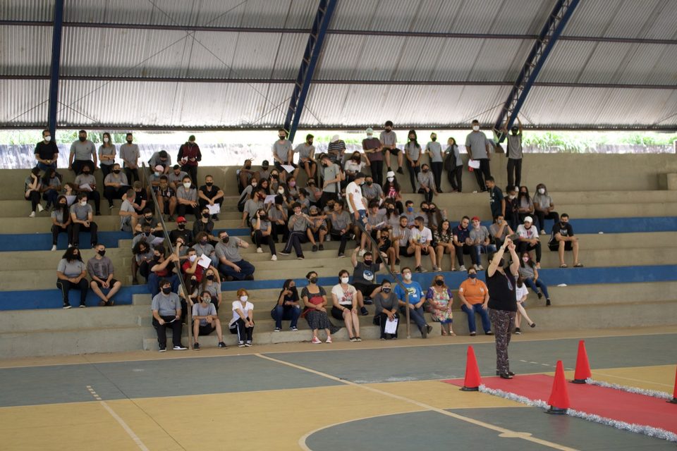 Estudantes na arquibancada; à frente, cones e um tapete vermelho no chão da quadra.