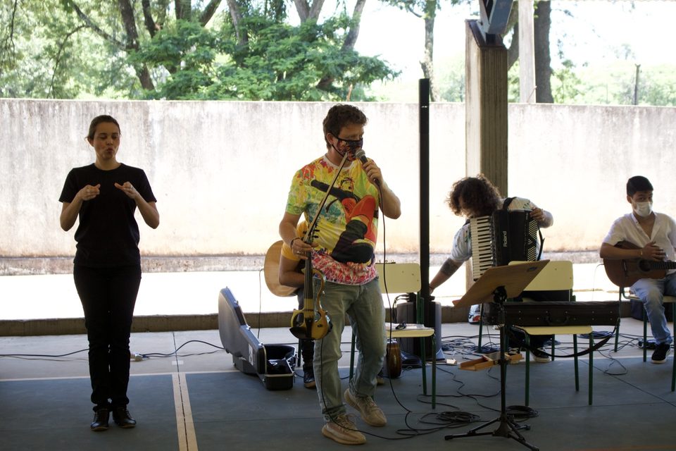 Uma mulher de preto gesticula ao lado do grupo, enquanto Roney Marczak toca.