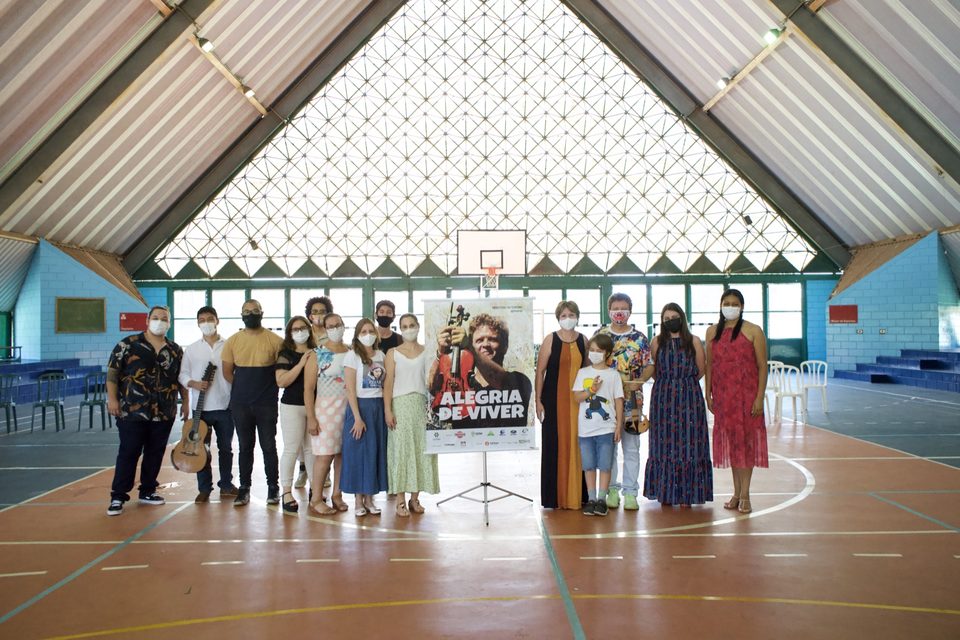 Músicos e professoras posam numa quadra coberta, ao lado do banner do disco.