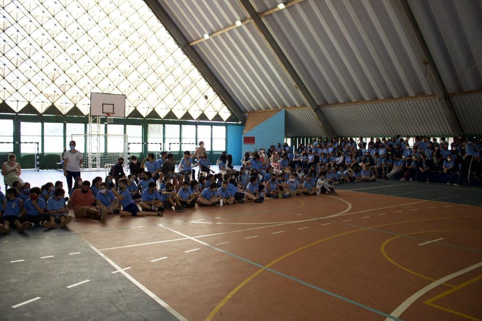 Estudantes de uniforme azul sentados no chão da quadra, de frente para os músicos.