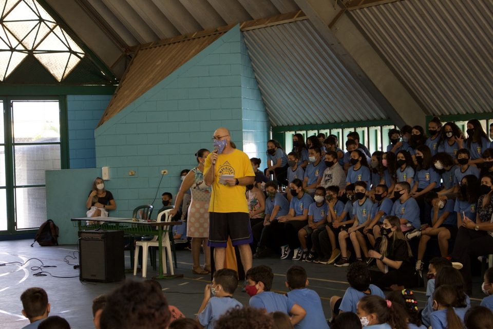 Um homem de camiseta amarela fala ao microfone diante dos estudantes sentados.