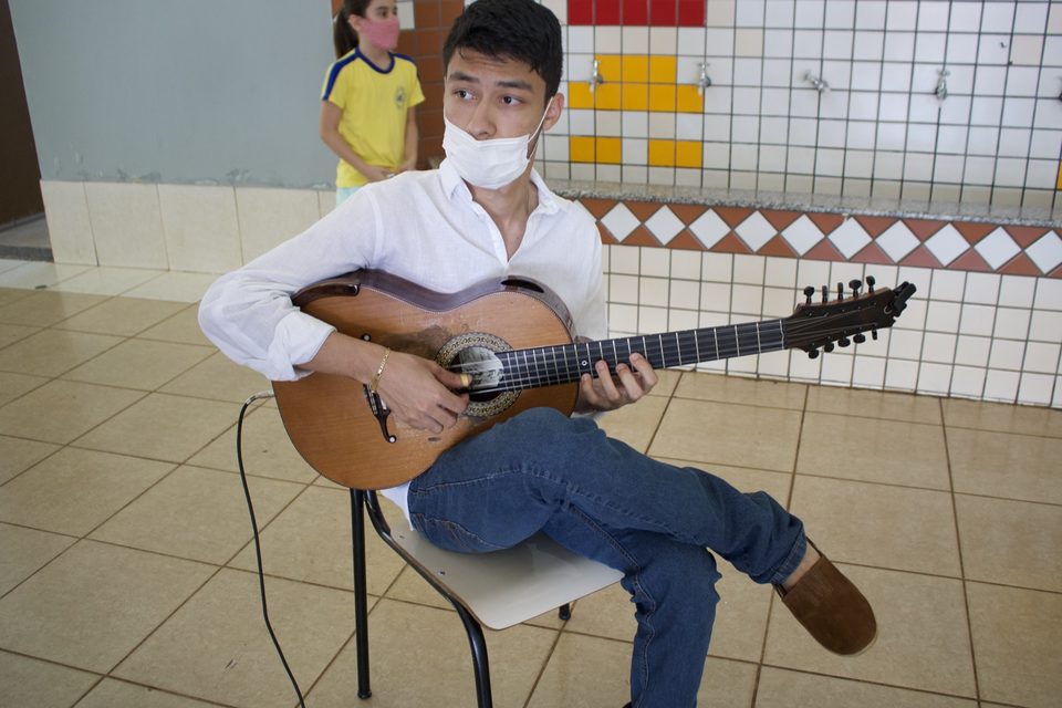 Um jovem de camisa branca dedilha um instrumento de cordas, sentado.