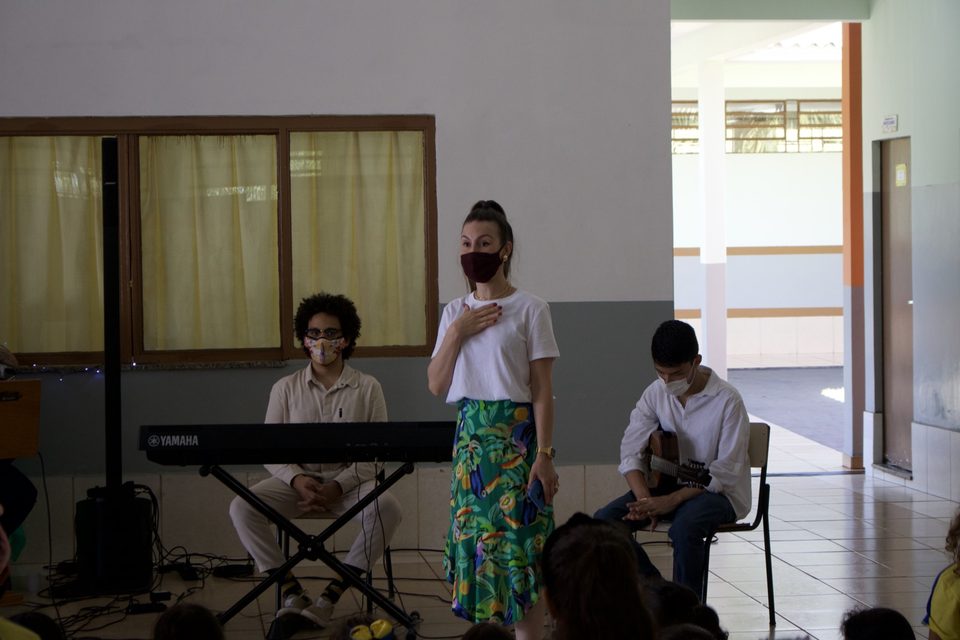 Uma mulher de saia verde gesticula entre o tecladista e o violonista.