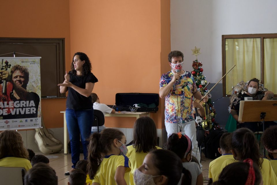 Roney Marczak toca violino diante das crianças, com o banner do disco à esquerda.