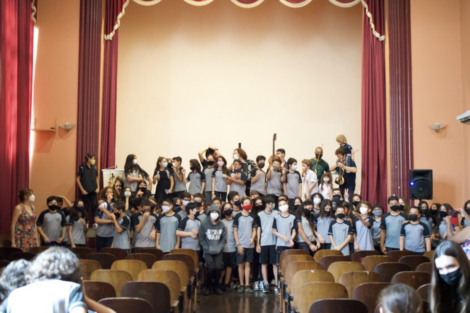 Dezenas de estudantes de uniforme cinza posam no palco do auditório, de braços erguidos.