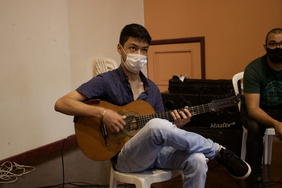 Um jovem de máscara toca violão, sentado.