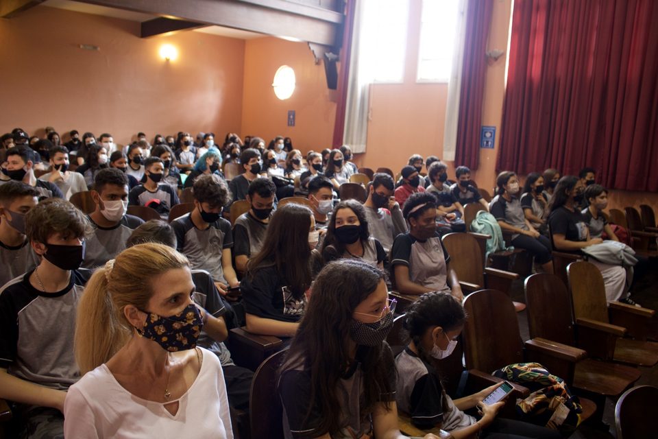 O auditório cheio de estudantes de máscara.