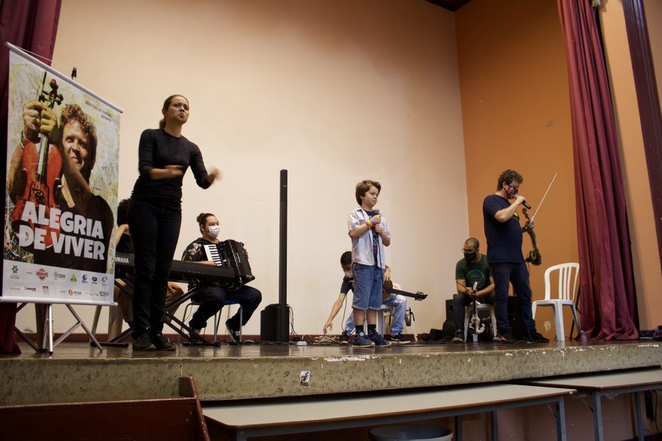Um menino canta ao microfone no palco, entre os músicos.