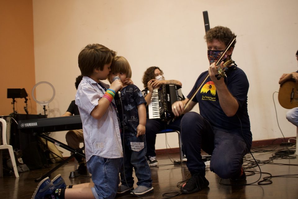 Roney Marczak, ajoelhado, toca violino ao lado de dois meninos; um deles segura o microfone. Ao fundo, a sanfona.