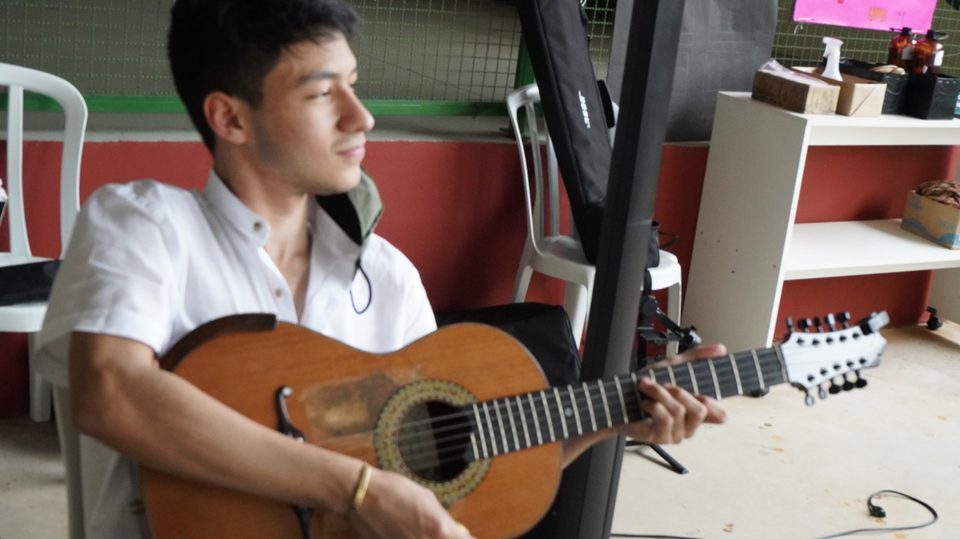 Um jovem músico de camisa branca toca violão, sentado.