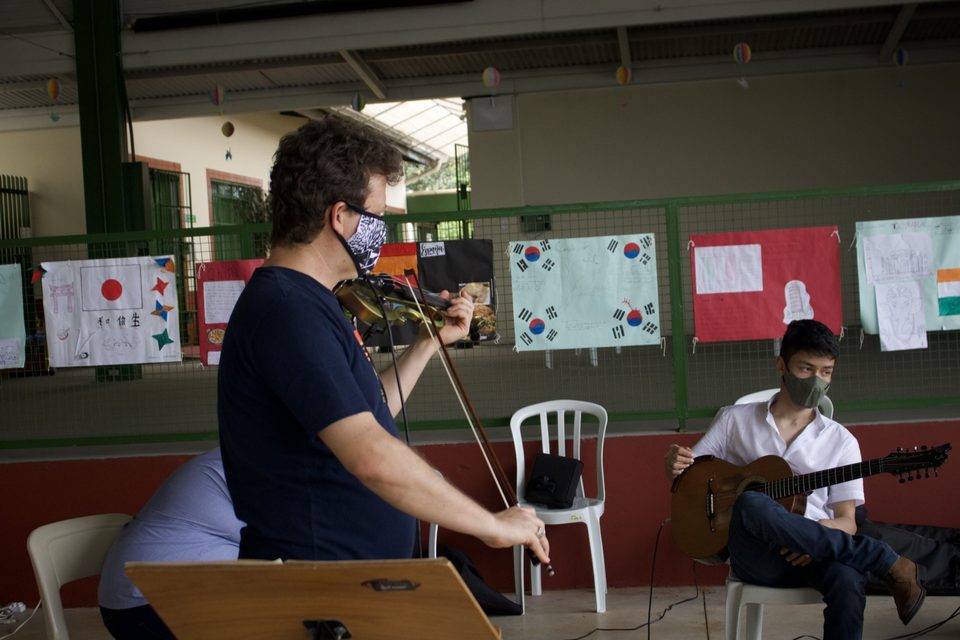 Roney Marczak toca violino; atrás, um violonista sentado e cartazes coloridos na parede.