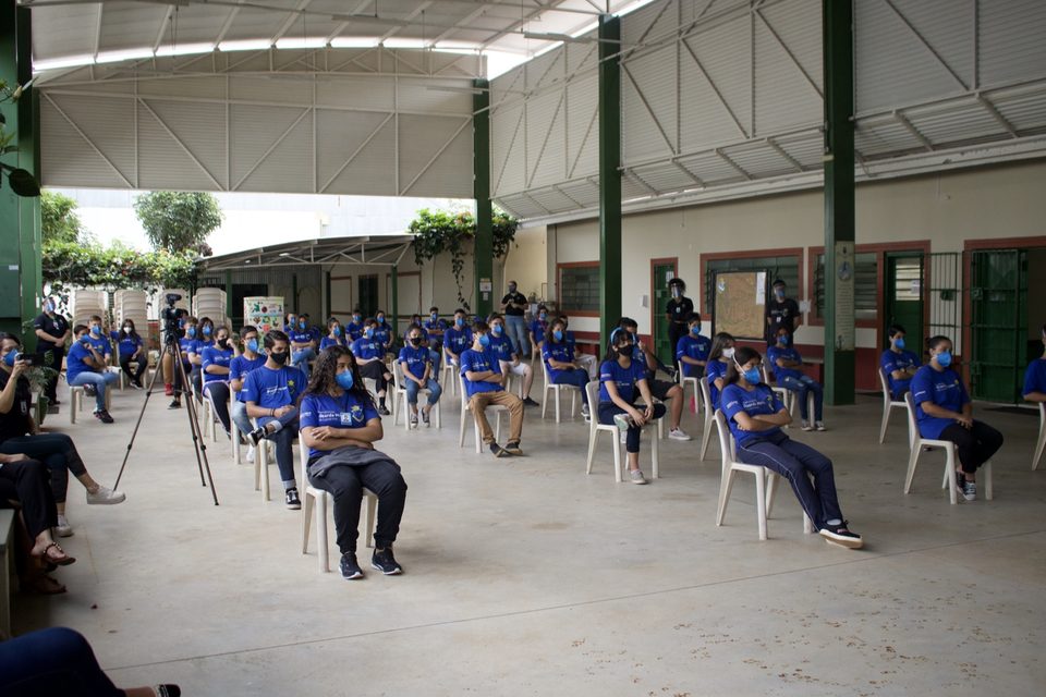 Jovens de camiseta azul sentados em cadeiras afastadas no pátio coberto.