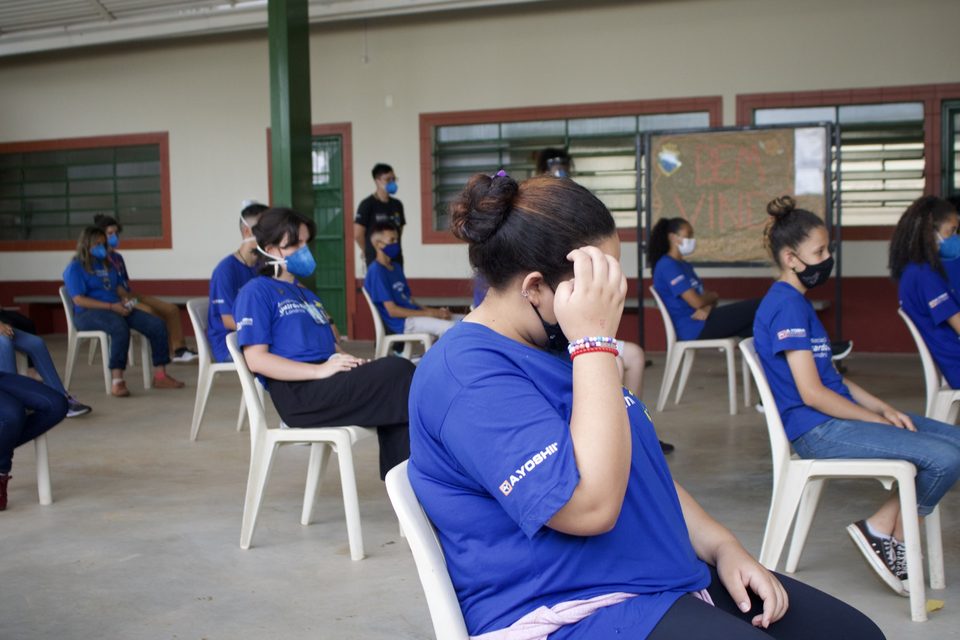 Jovens de camiseta azul e máscara ouvem a apresentação, sentados.