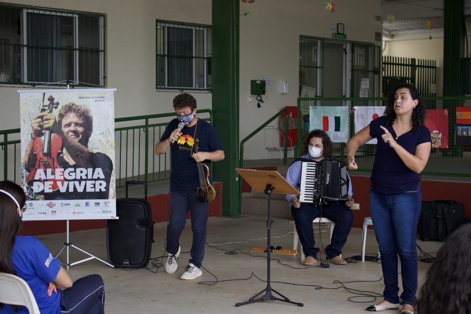 Ao lado do banner do disco, uma mulher gesticula enquanto os músicos tocam.