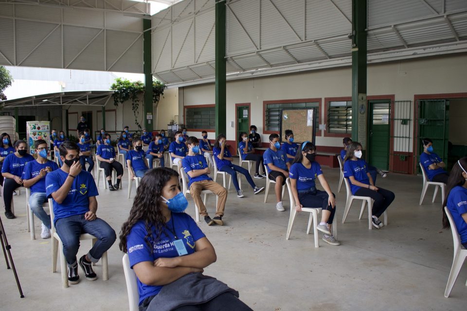 Uma jovem de camiseta azul ouve de braços cruzados; atrás, os colegas.