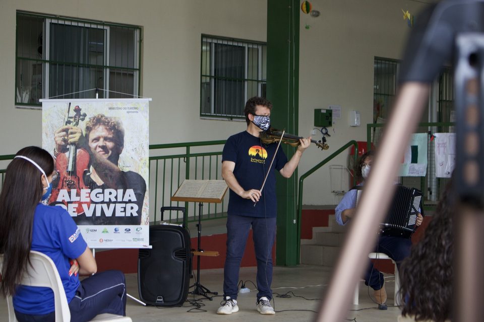 Roney Marczak toca violino ao lado do banner; em primeiro plano, uma jovem assiste.