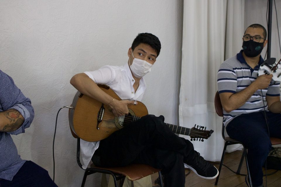 Um jovem toca violão; ao lado, um músico segura um violino.