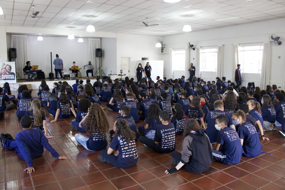 Estudantes sentados no chão do salão, de frente para o palco.