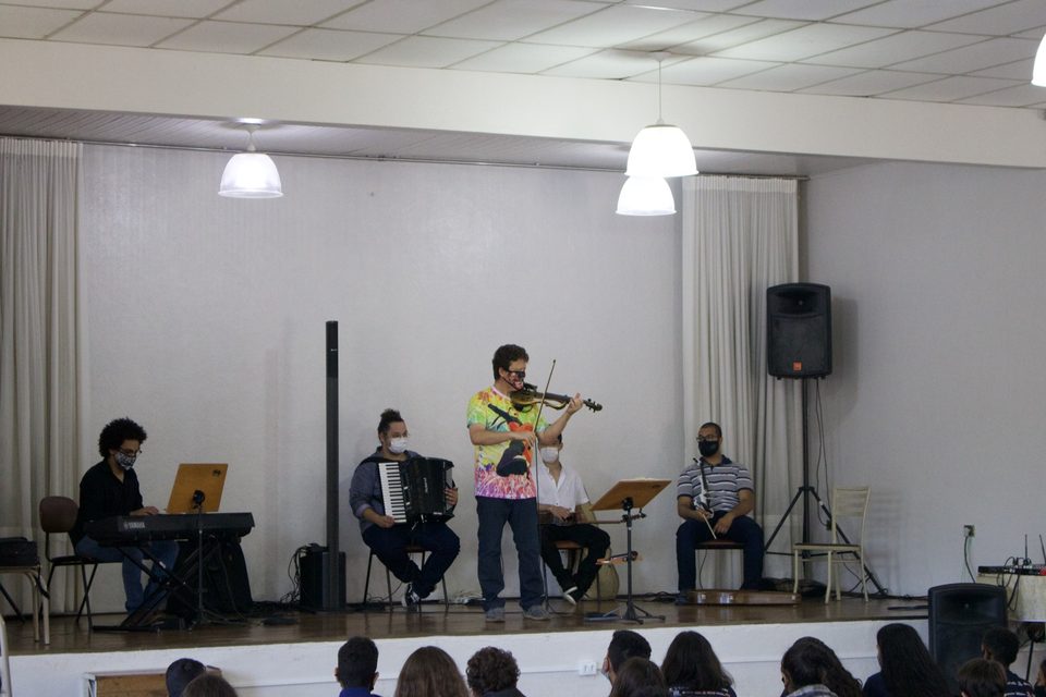 Roney Marczak toca violino ao centro do palco, entre os músicos.