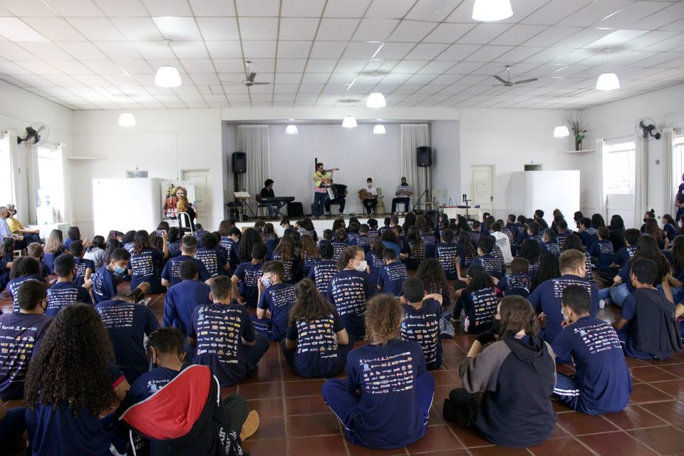 O salão cheio de estudantes sentados no chão, com o palco ao fundo.