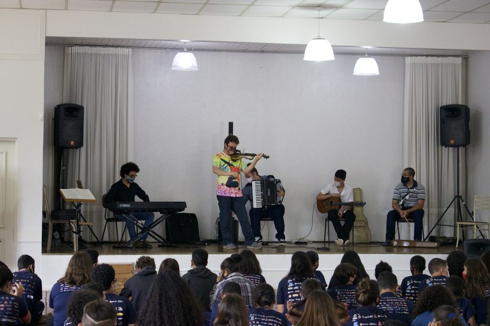 Roney Marczak toca violino ao centro; estudantes assistem em primeiro plano.