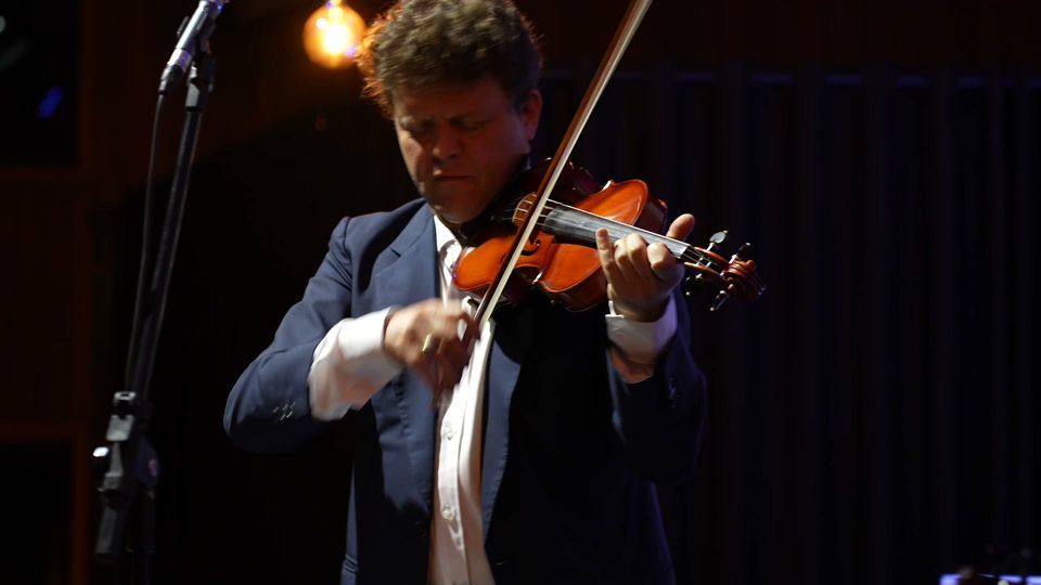 Roney Marczak, de terno escuro, toca violino de pé junto ao microfone.
