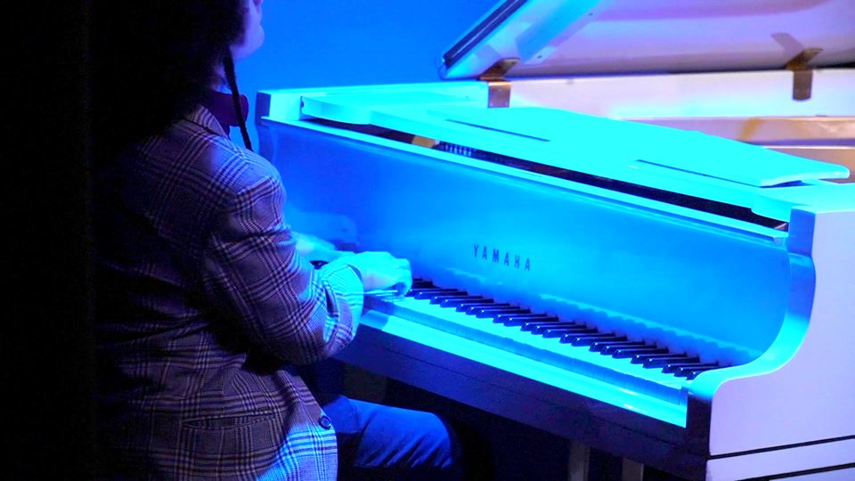 Um pianista toca o piano de cauda branco, iluminado de azul.