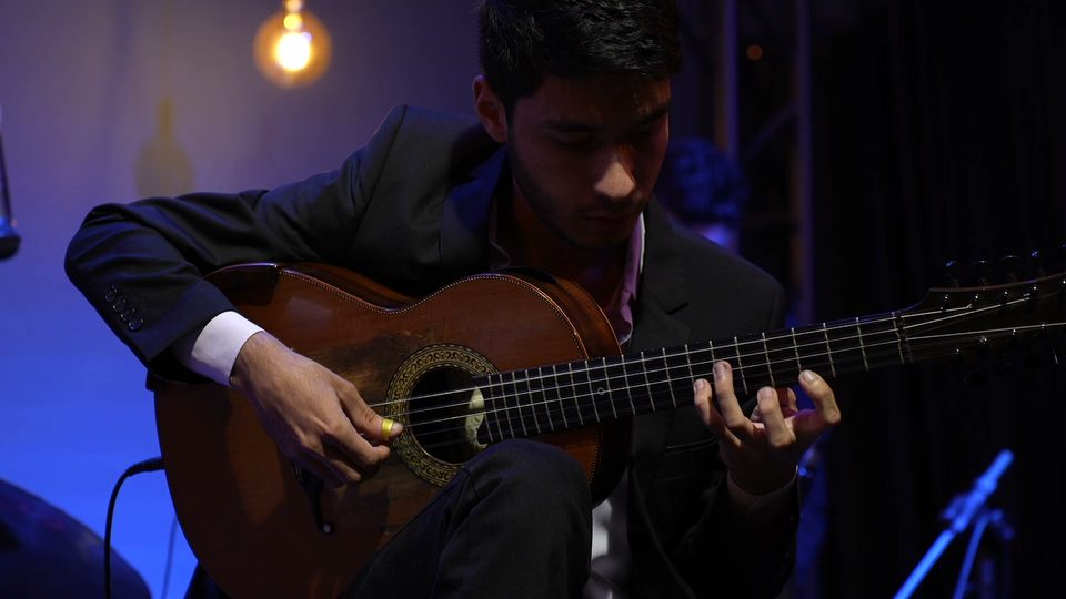 Um jovem músico de terno toca viola caipira, sentado, sob luz azul.