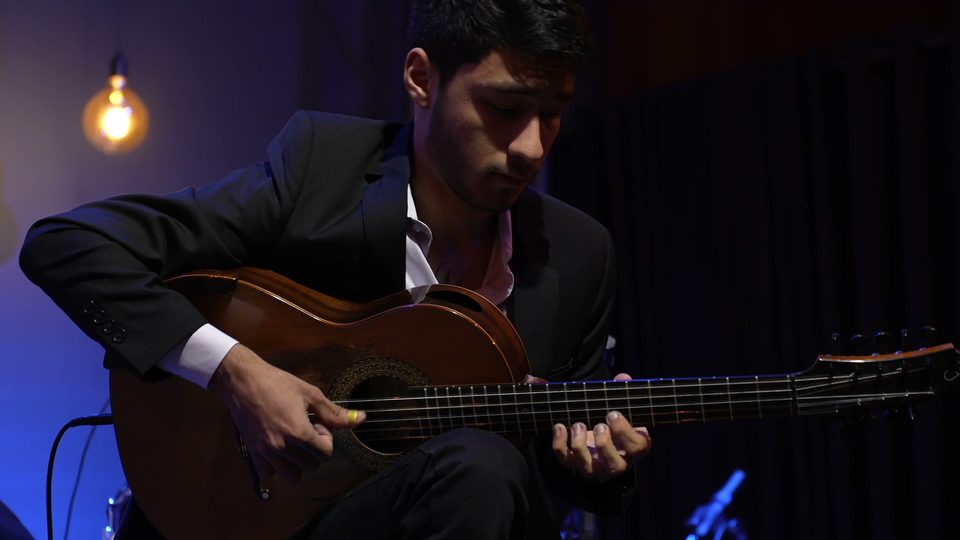 O mesmo músico, de perfil, dedilha a viola; ao fundo, uma lâmpada acesa.