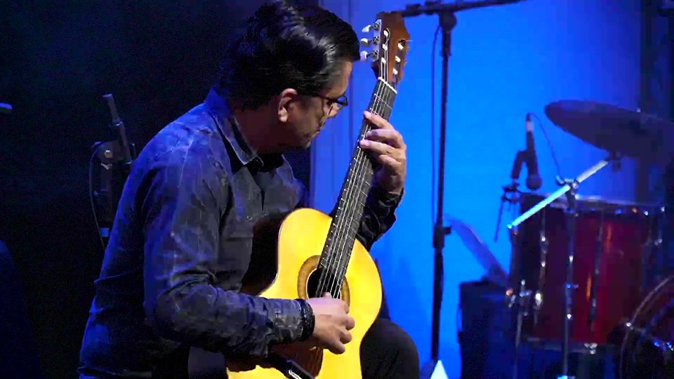 Um violonista de óculos toca violão de tampo claro, ao lado da bateria.