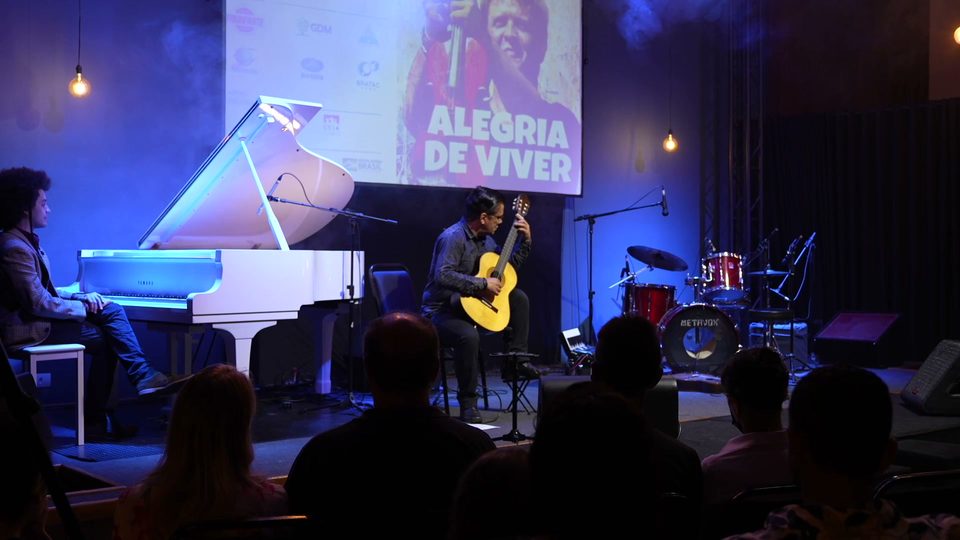 Vista ampla do palco: o piano de cauda branco, o violonista ao centro e a plateia em primeiro plano.