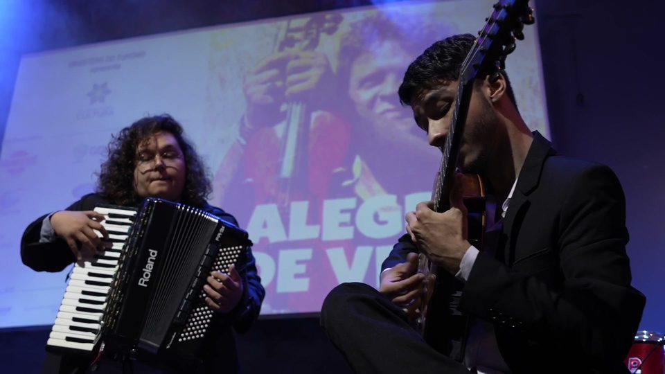 Sanfona e um instrumento de cordas tocados lado a lado, diante do telão.