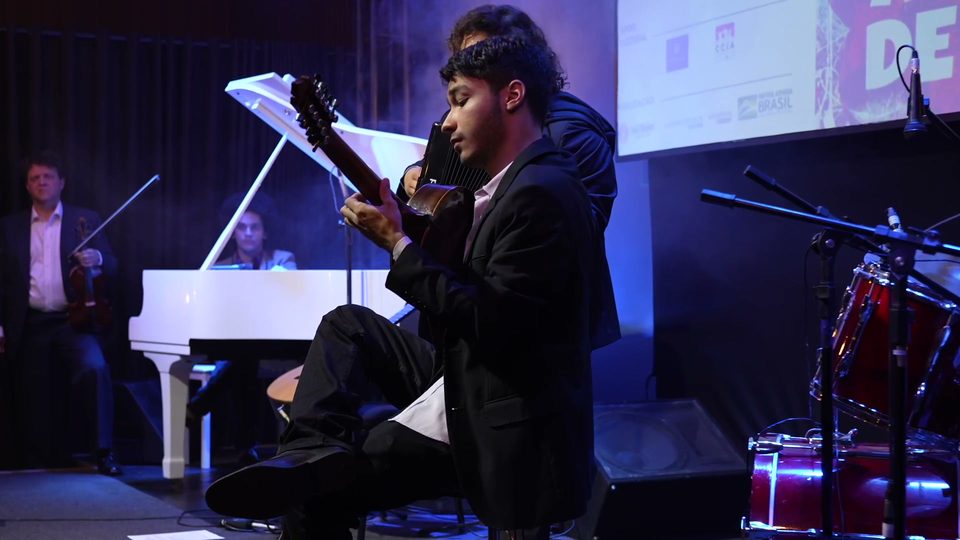 Um jovem músico toca sentado na beira do palco; ao fundo, o piano branco.