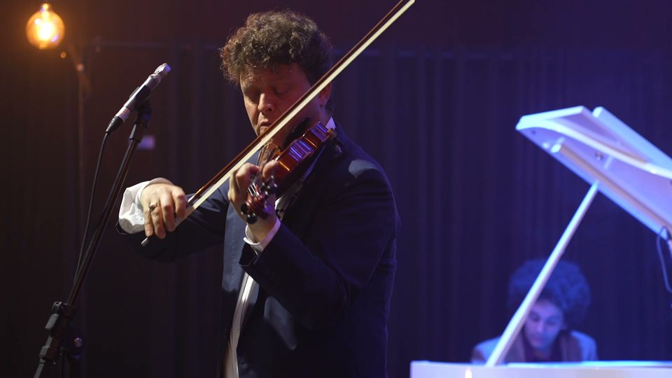 Roney Marczak toca violino ao lado da tampa aberta do piano branco.