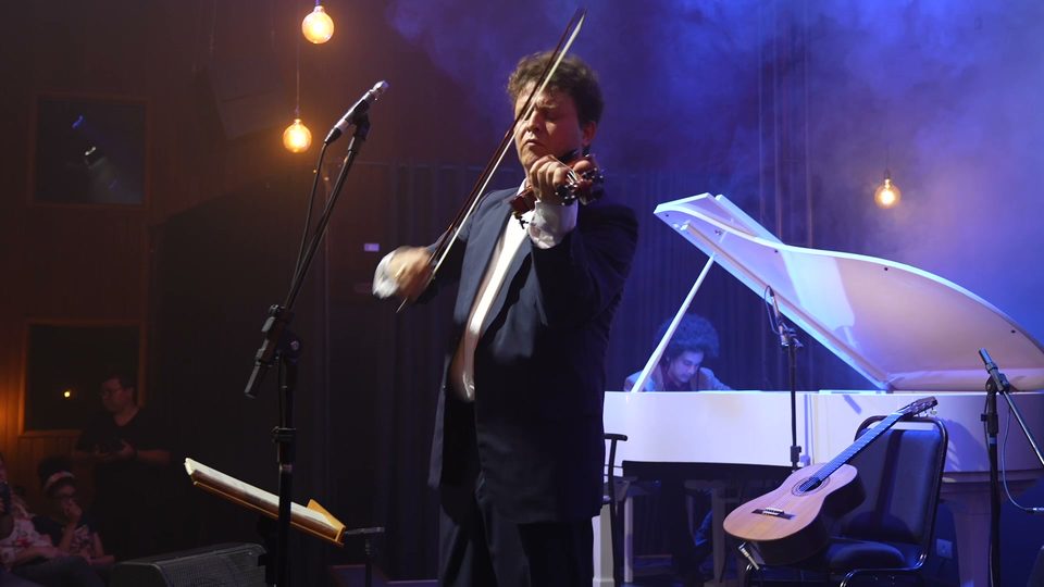 Roney Marczak toca violino em meio à fumaça cênica, com o piano branco atrás.