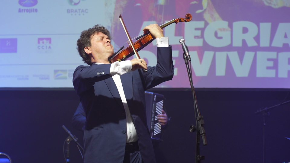 Roney Marczak toca violino de braços abertos diante do telão com o título Alegria de Viver.