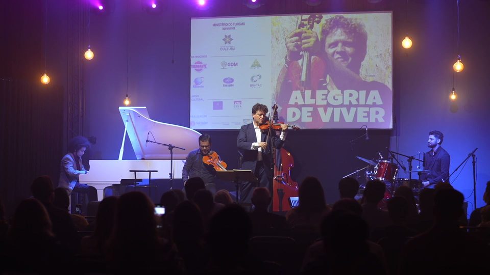 O grupo no palco: piano branco, contrabaixo, violino e bateria.