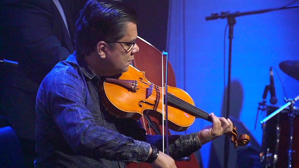 Um músico de óculos toca um instrumento de arco, iluminado de azul.