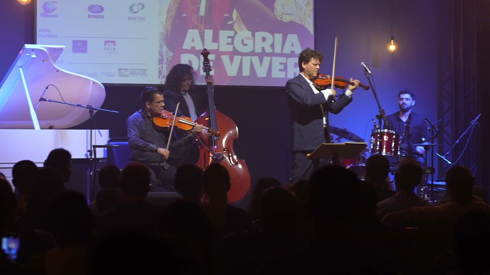O palco com o piano branco, o contrabaixo e os violinos, diante do telão do disco.