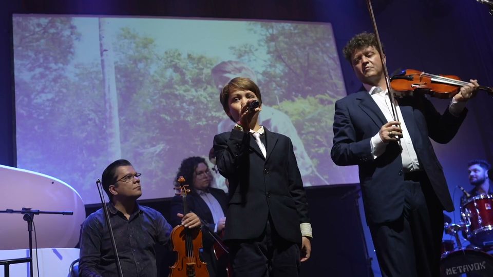 Um menino de terno canta ao microfone ao lado de Roney Marczak, que segura o violino.