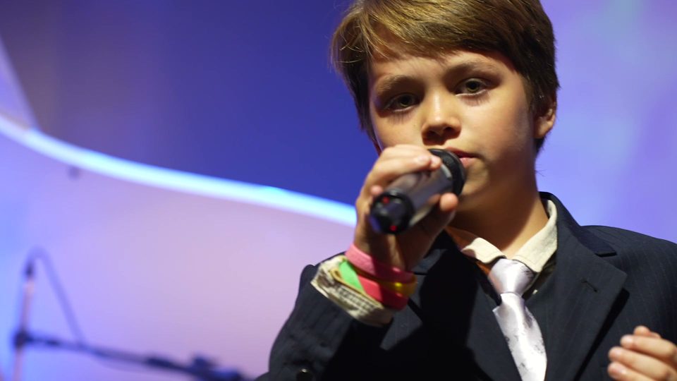 O menino canta ao microfone, em primeiro plano.