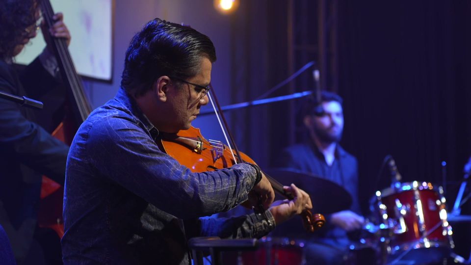 Um músico de óculos toca um instrumento de arco; atrás, a bateria.
