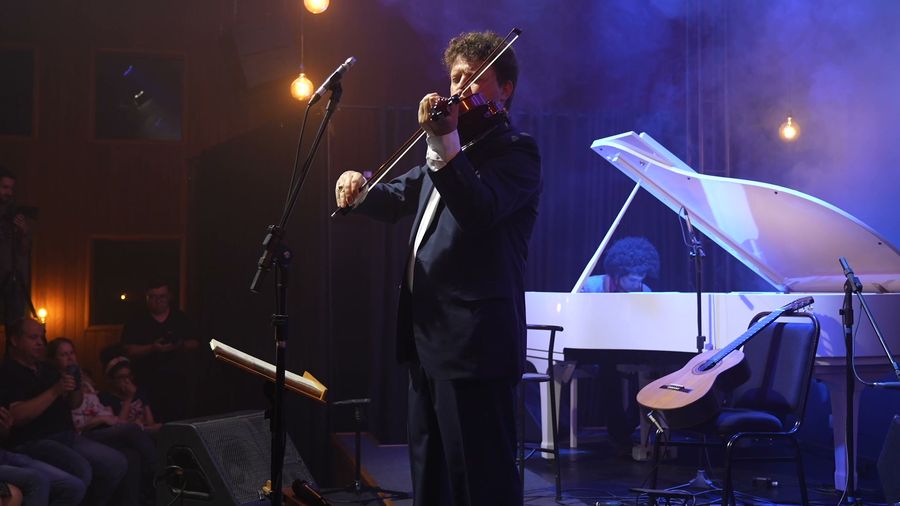 Roney Marczak, de terno preto, toca violino em um palco iluminado de azul; ao fundo, um piano de cauda branco com pianista sentado.