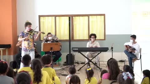 Vídeo: Roney Marczak toca violino no salão, com crianças de uniforme amarelo sentadas à frente.