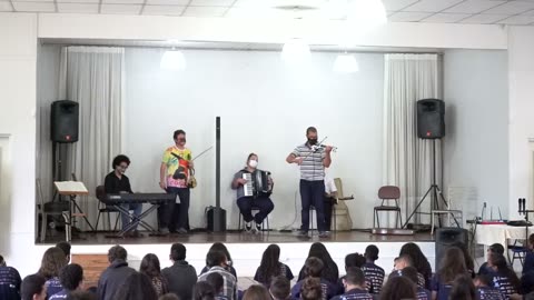 Vídeo: o grupo toca no palco do salão, com os estudantes sentados em primeiro plano.